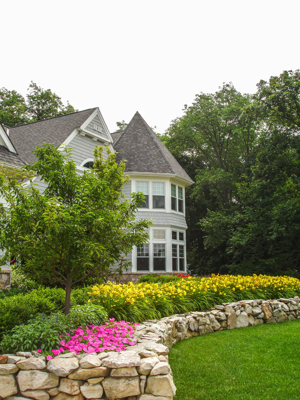 Estate house with lawn, stone wall, yellow and pink seasonal color, trees, and layered garden planting